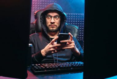 IT-Sicherheit und Cybersecurity: Ein Mann mit Brille und Kapuzenpullover sitzt vor zwei Monitoren in einem abgedunkelten Raum mit schalldämmenden Wänden. Er hält ein Smartphone und blickt überrascht auf den Bildschirm. Die Szene mit blauen und roten Lichtakzenten vermittelt eine Hacking- oder Cyberangriff-Atmosphäre.